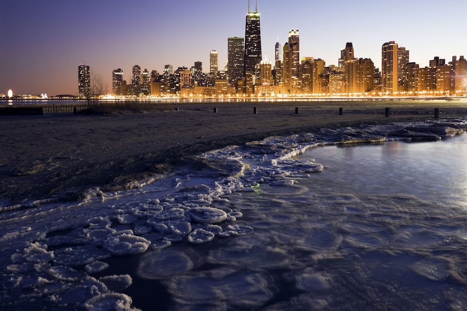A frozen river with ice formations, a city skyline with illuminated buildings, and a bridge in the background.