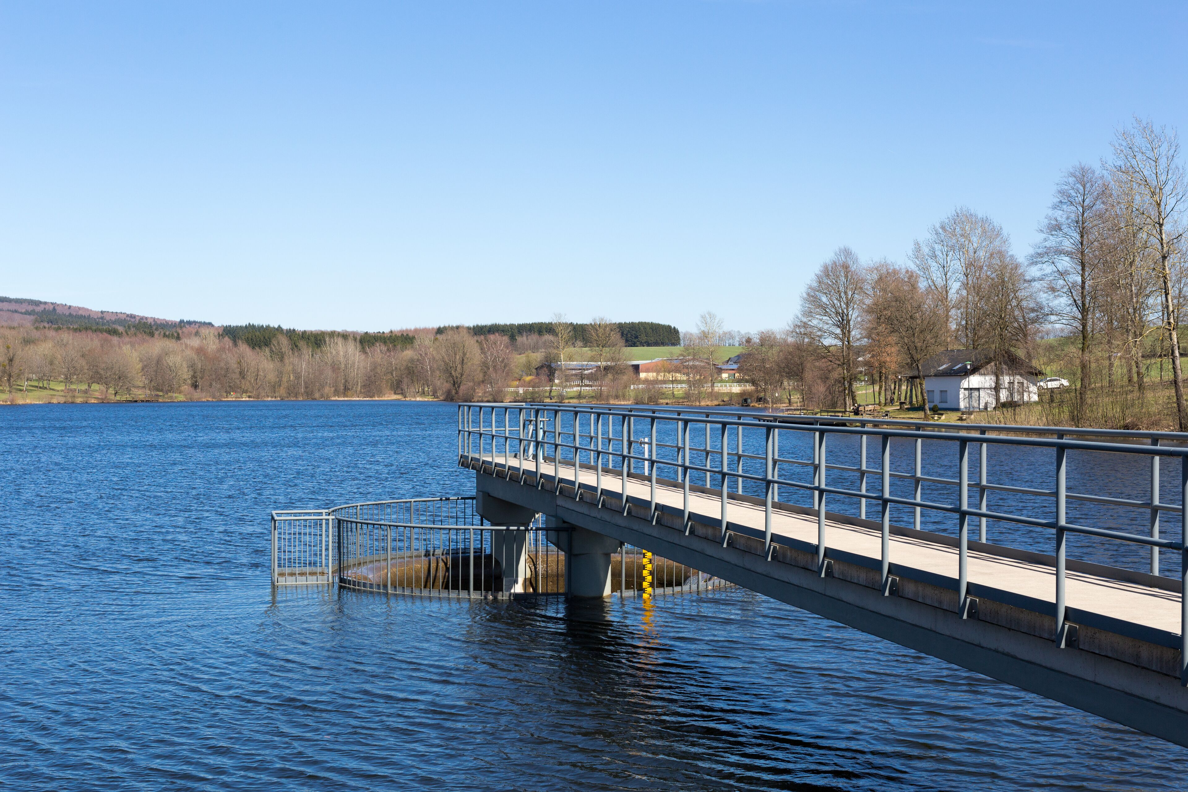 A bridge over a calm lake with a clear sky, surrounded by trees and buildings.