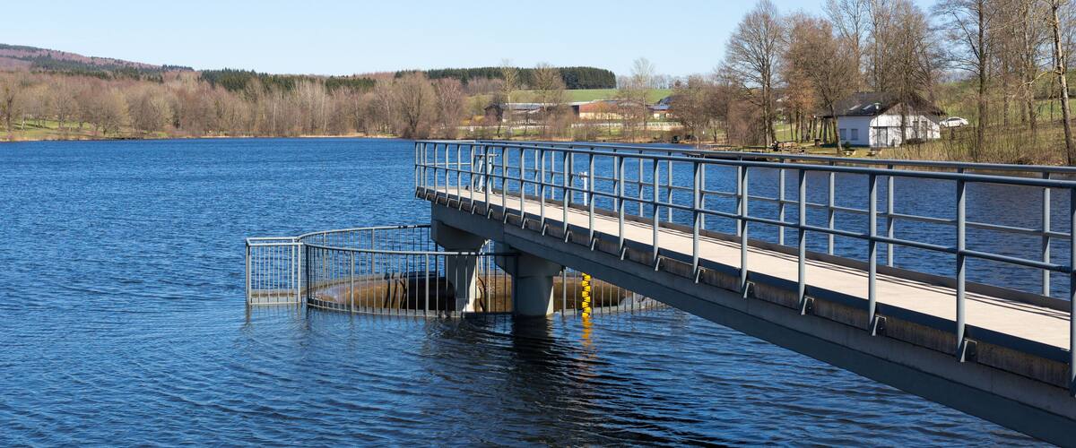 A bridge over a calm lake with a clear sky, surrounded by trees and buildings.