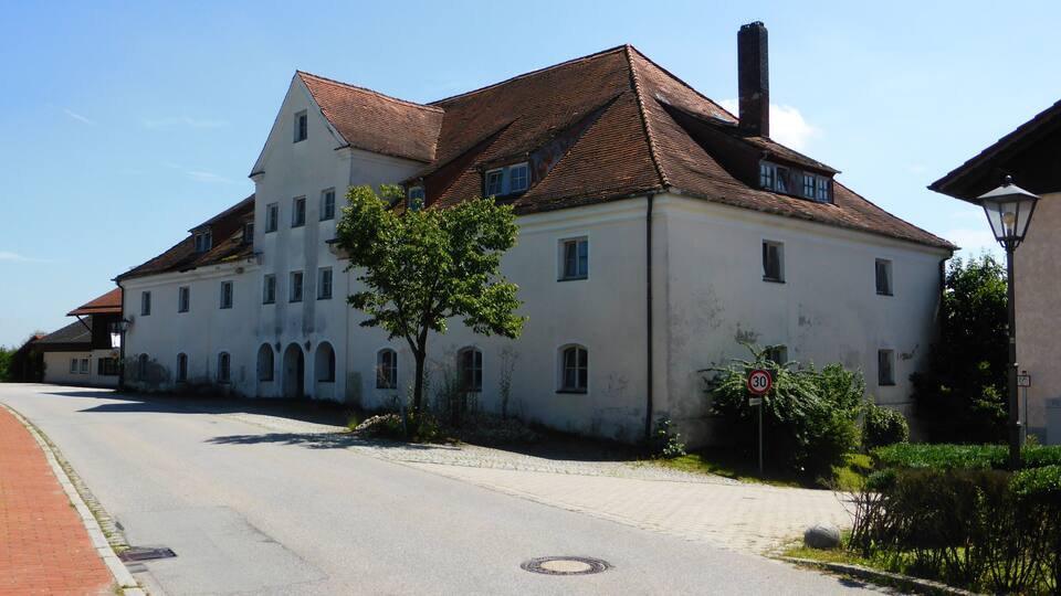 A large white building with a red-tiled roof, a tree in front, and a cobblestone street.