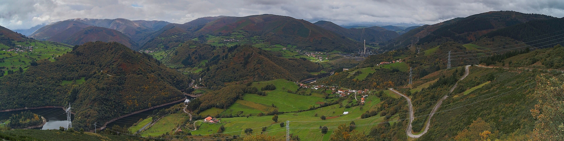 Paisaje montañoso con un camino sinuoso, un pequeño pueblo y una torre de líneas eléctricas.