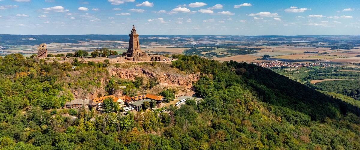 A historic castle on a hill with a tower, surrounded by dense forest and a small village below.