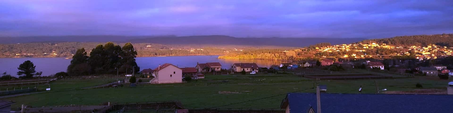 A coastal town at dusk with a lake, houses, and a mountain in the background.