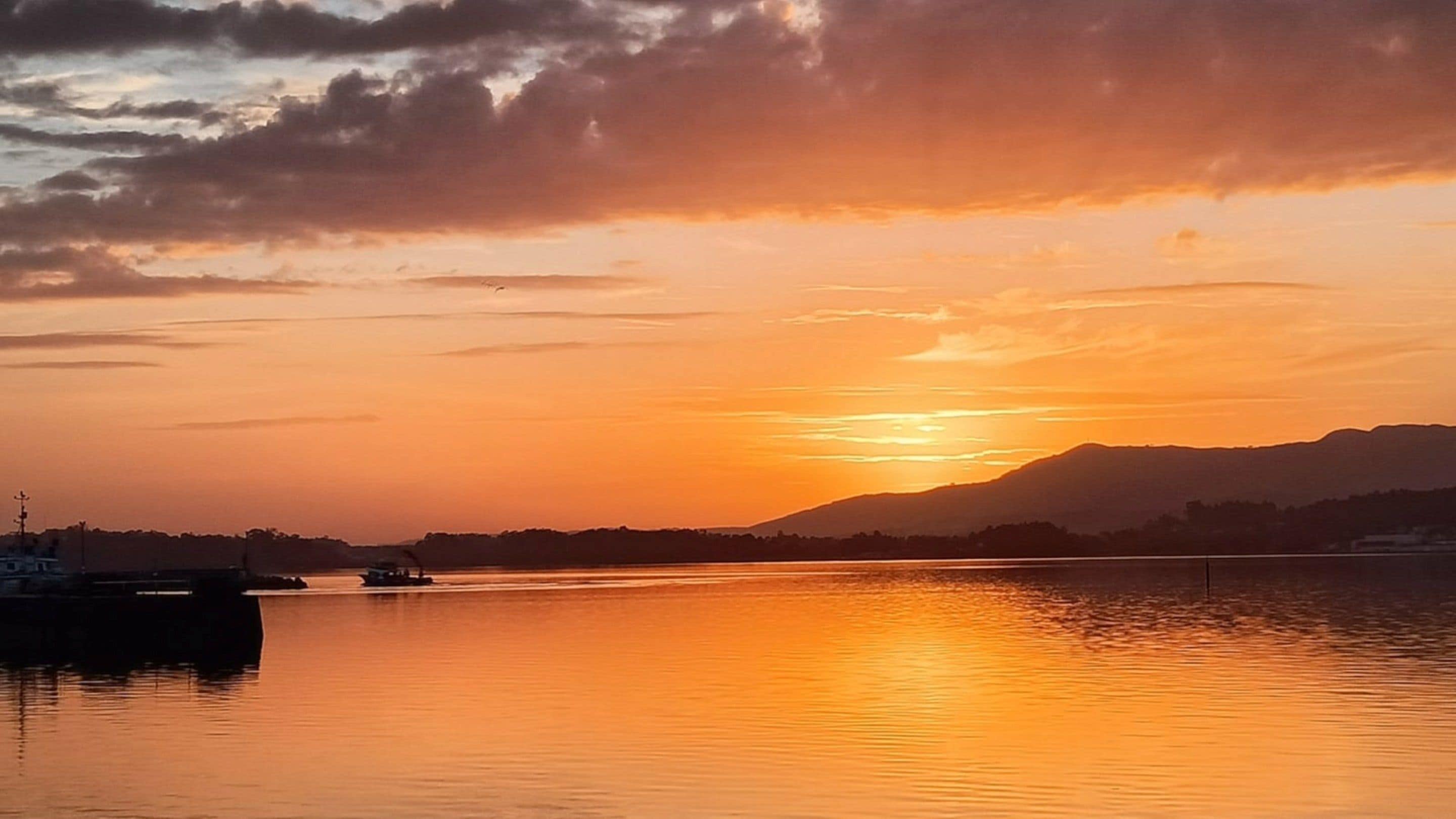 A sunset over a calm lake with a boat docked on the left and mountains in the background.
