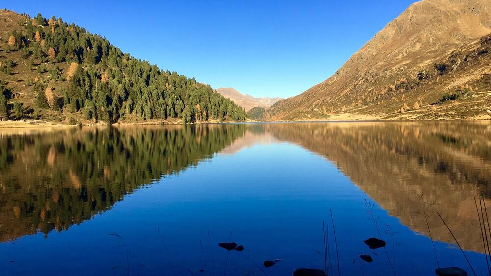 Ein ruhiger See mit klaren Spiegelungen, umgeben von bewaldeten Hügeln und Bergen.