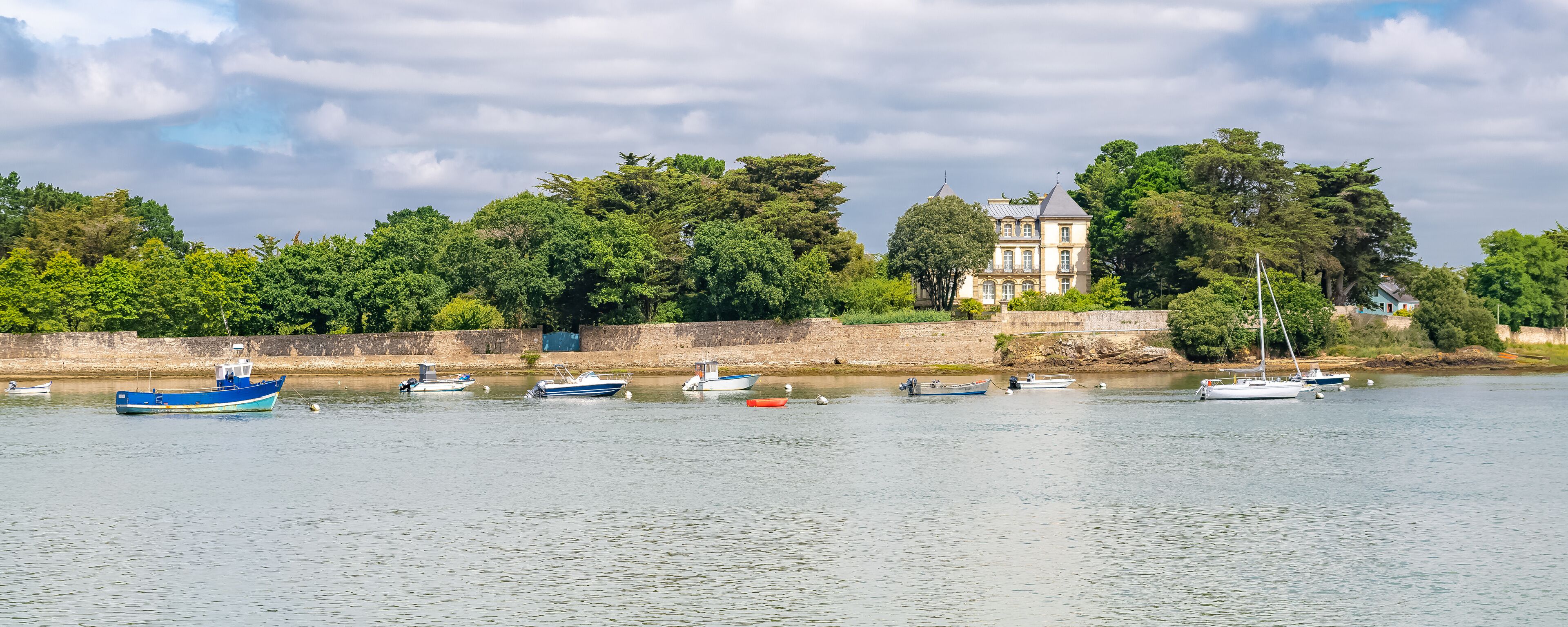 A calm river with several boats docked along the shore, a large tree-lined building, and a stone wall.