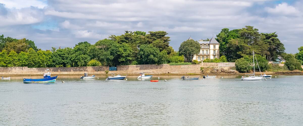 A calm river with several boats docked along the shore, a large tree-lined building, and a stone wall.