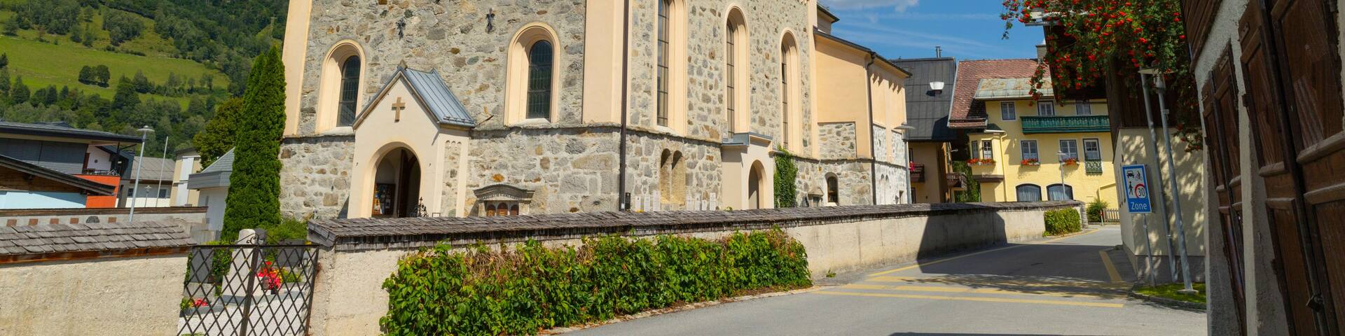 A church with a stone facade and a tall steeple, set against a backdrop of a clear blue sky and green hills.