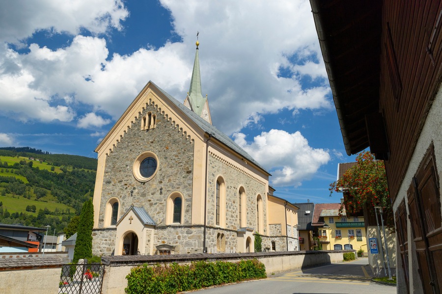 A church with a stone facade and a tall steeple, set against a backdrop of a clear blue sky and green hills.
