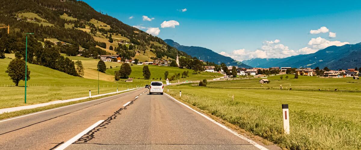 A rural road with a white van, surrounded by green fields, trees, and a mountain range in the background.