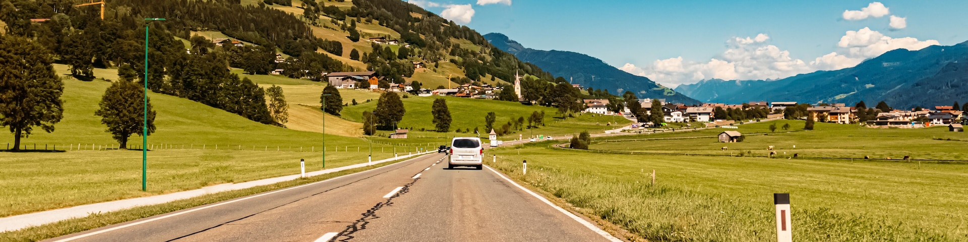 A rural road with a white van, surrounded by green fields, trees, and a mountain range in the background.