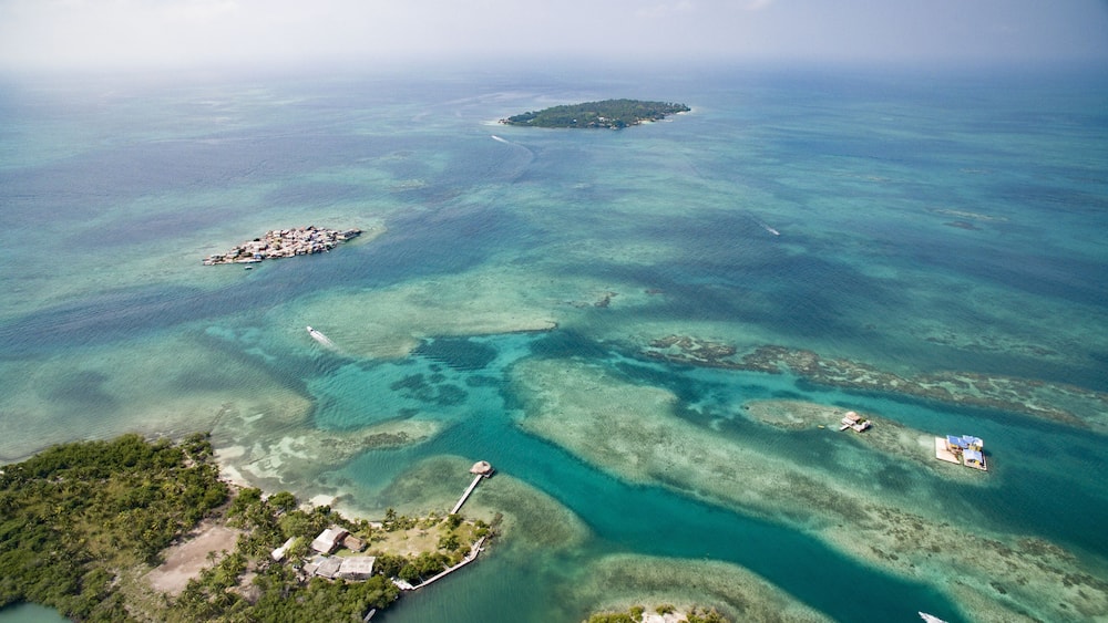An aerial view of a tropical island with clear turquoise waters, a small settlement, and a distant larger island.