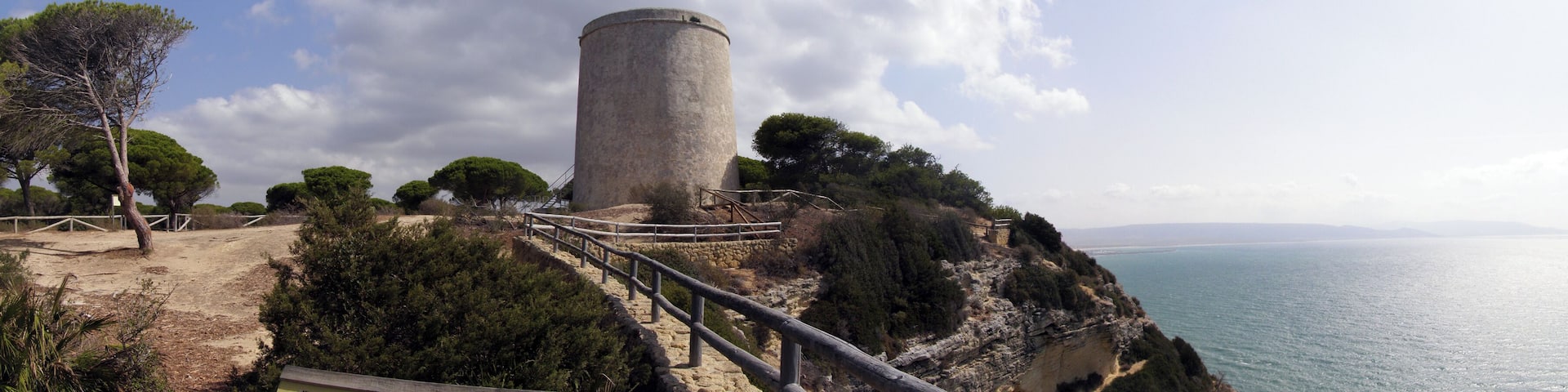 A coastal lighthouse on a cliff with a wooden walkway and a view of the sea.