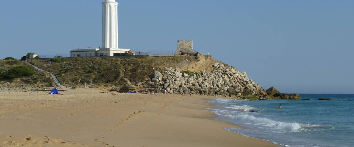 A white lighthouse on a rocky cliff by a sandy beach with waves crashing onto the shore.
