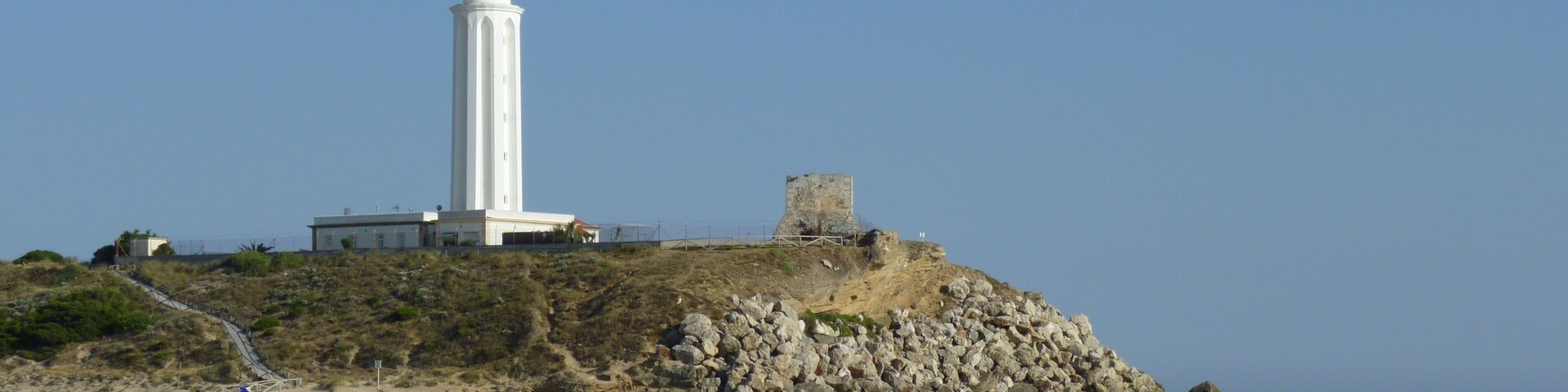 A white lighthouse on a rocky cliff by a sandy beach with waves crashing onto the shore.