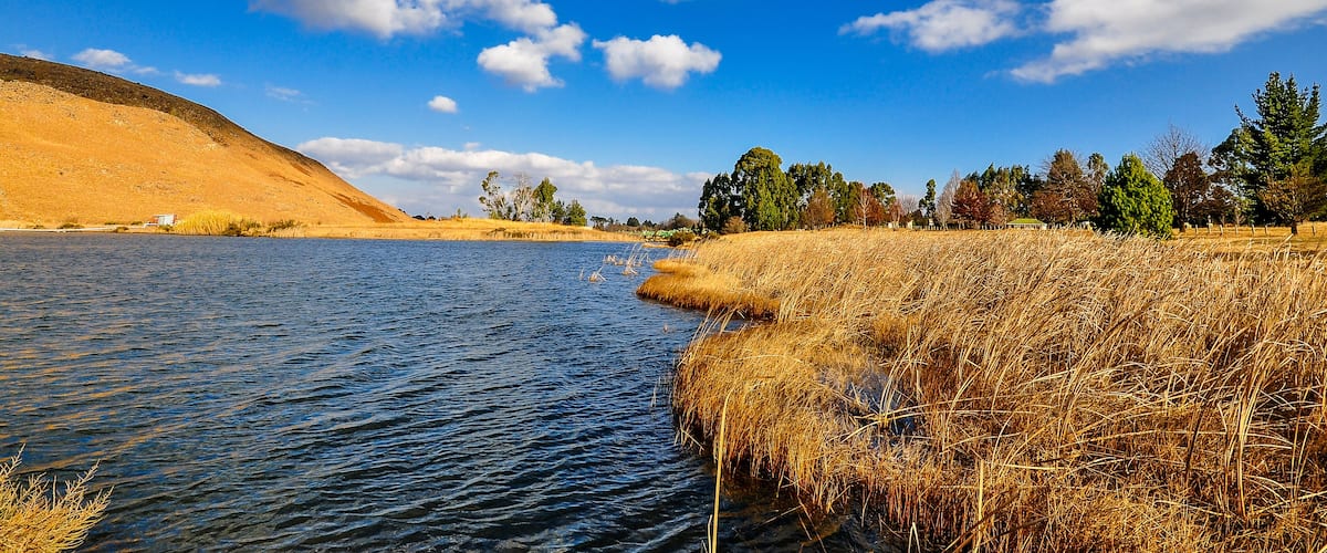 A river with dry grass on the banks, a hill in the background, and a clear blue sky with clouds.