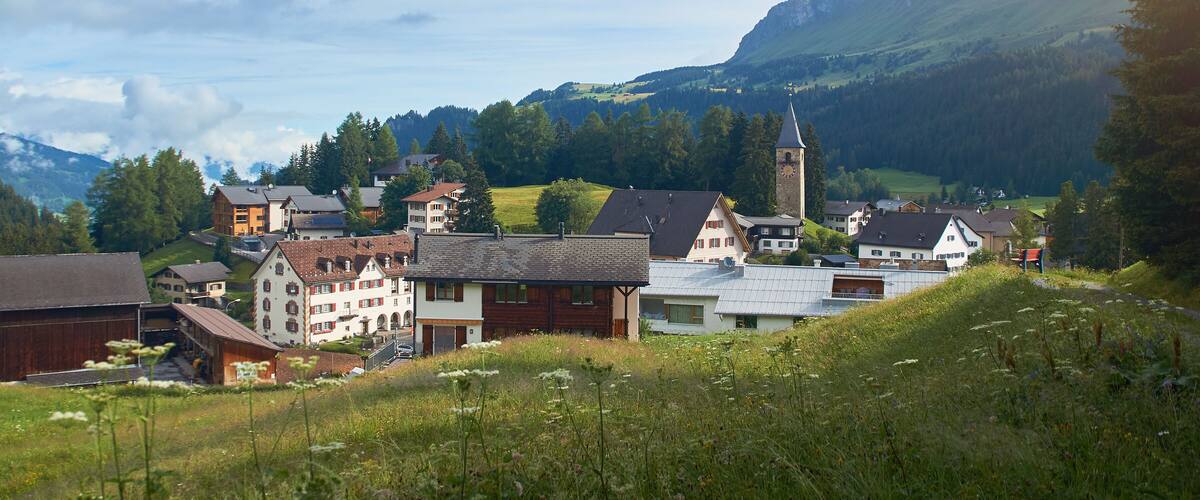 A village with traditional houses, a church with a clock tower, and mountains in the background.