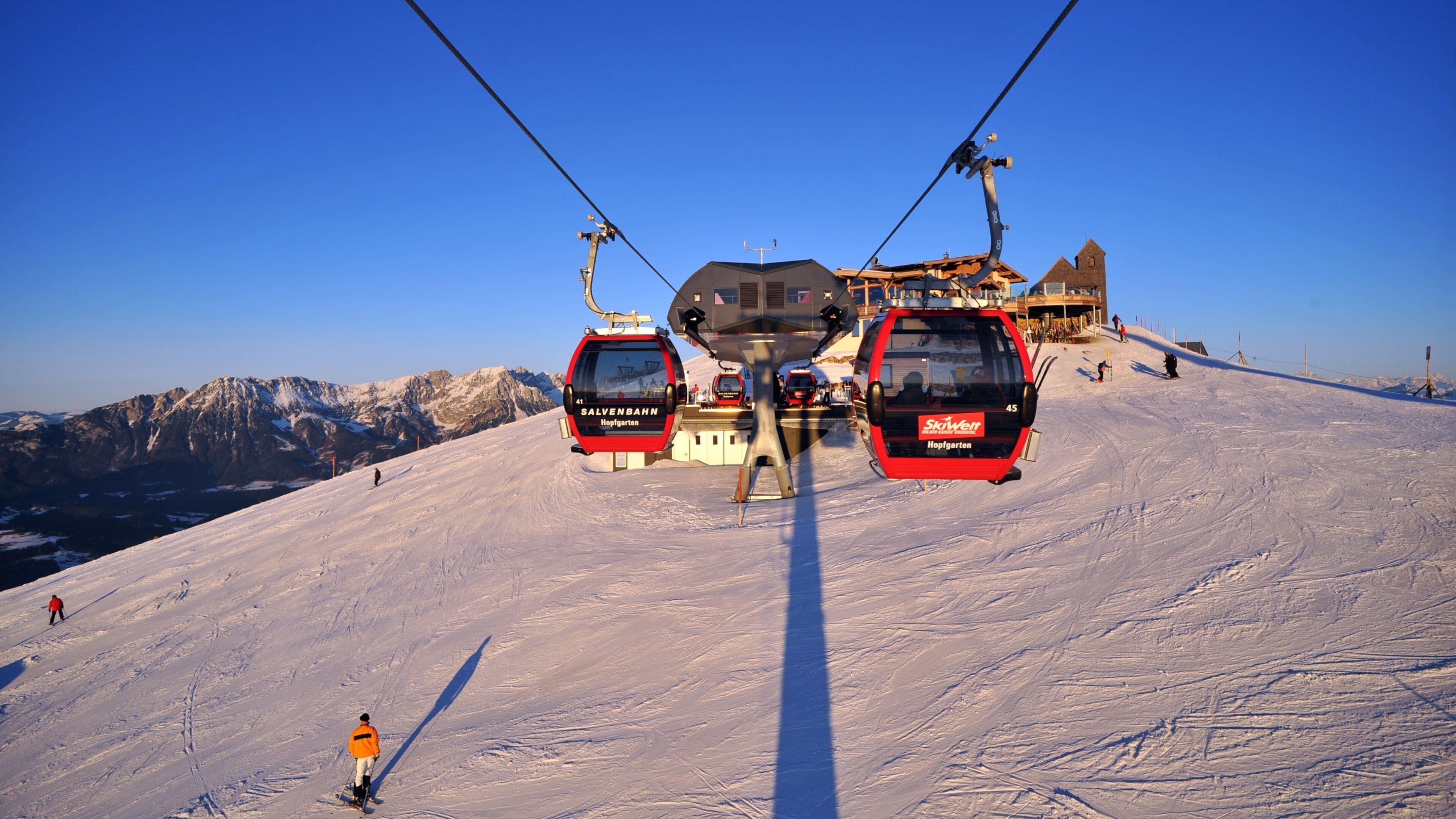 A ski lift with two red gondolas, a mountain backdrop, and a building with a tower.