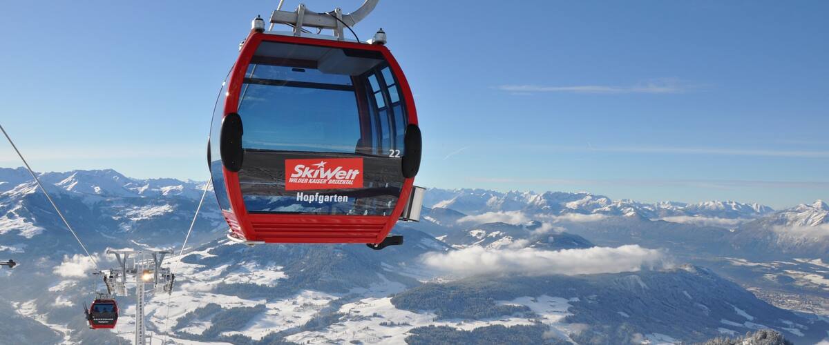 A red ski lift chair suspended over a snowy mountain landscape with snow-covered trees and distant peaks.