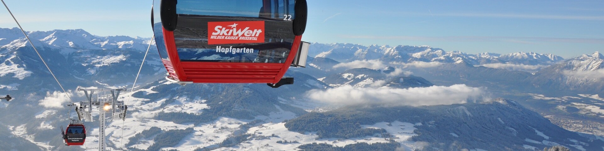 A red ski lift chair suspended over a snowy mountain landscape with snow-covered trees and distant peaks.