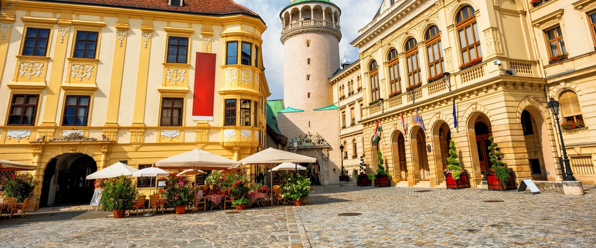 A cobblestone square with historic buildings, a prominent tower with a green dome, and outdoor seating with umbrellas.