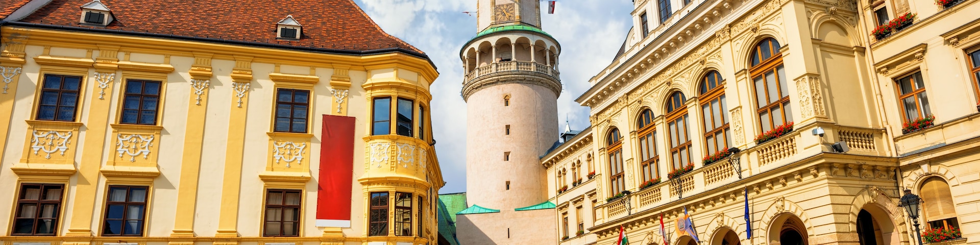 A cobblestone square with historic buildings, a prominent tower with a green dome, and outdoor seating with umbrellas.