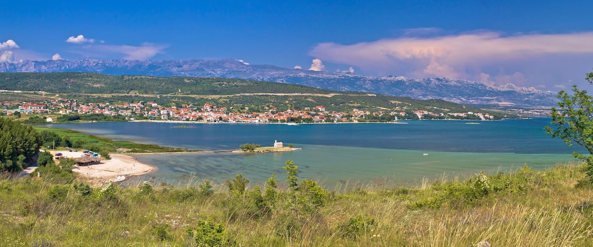 A coastal town with a clear view of the sea, mountains in the background, and a small island with a building.