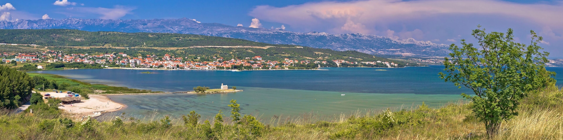 A coastal town with a clear view of the sea, mountains in the background, and a small island with a building.