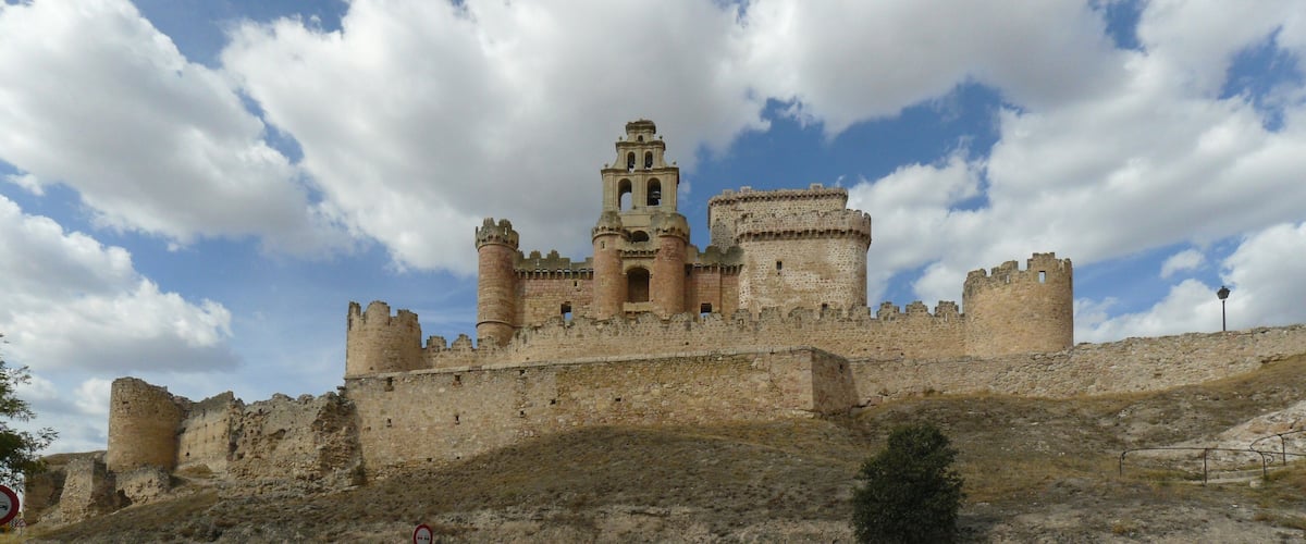 A historic castle with multiple towers and a prominent bell tower, surrounded by a stone wall and set against a partly cloudy sky.