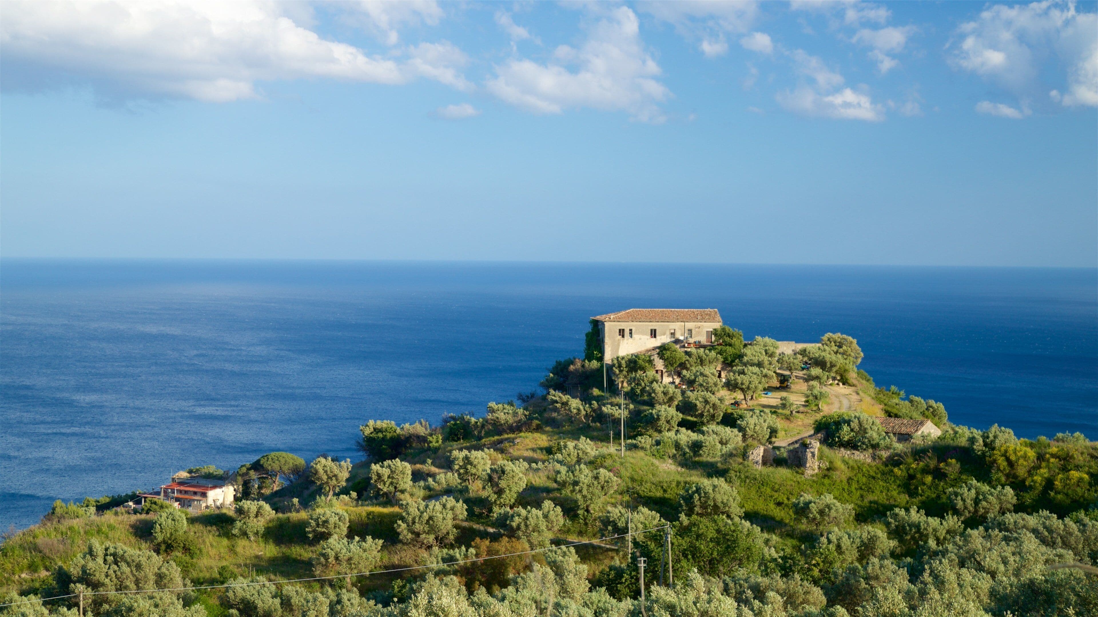 A coastal landscape with a prominent building on a hill, surrounded by olive trees and a clear view of the sea.