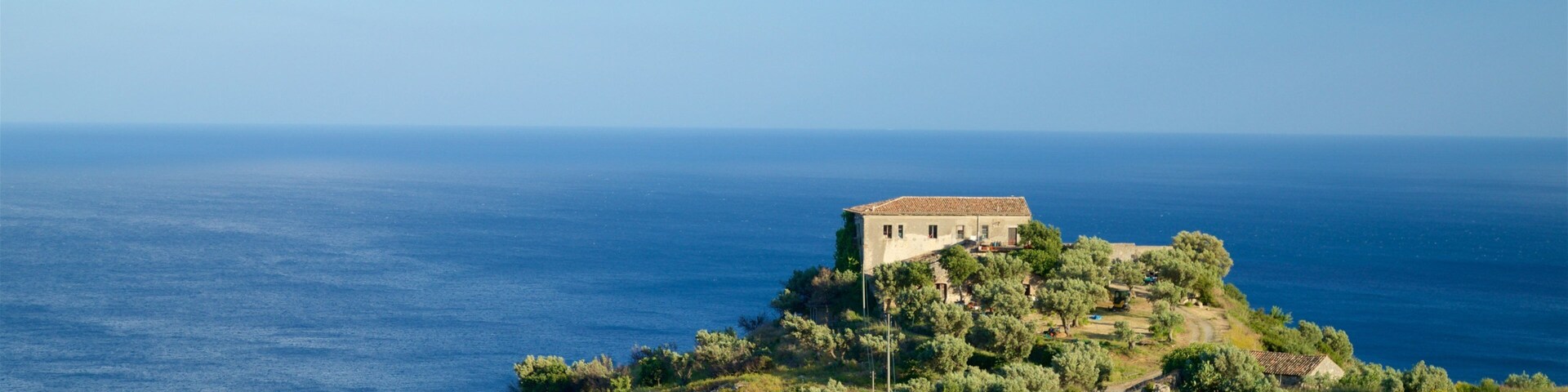 Un paisaje costero con un edificio destacado en una colina, rodeado de olivos y una vista despejada del mar.