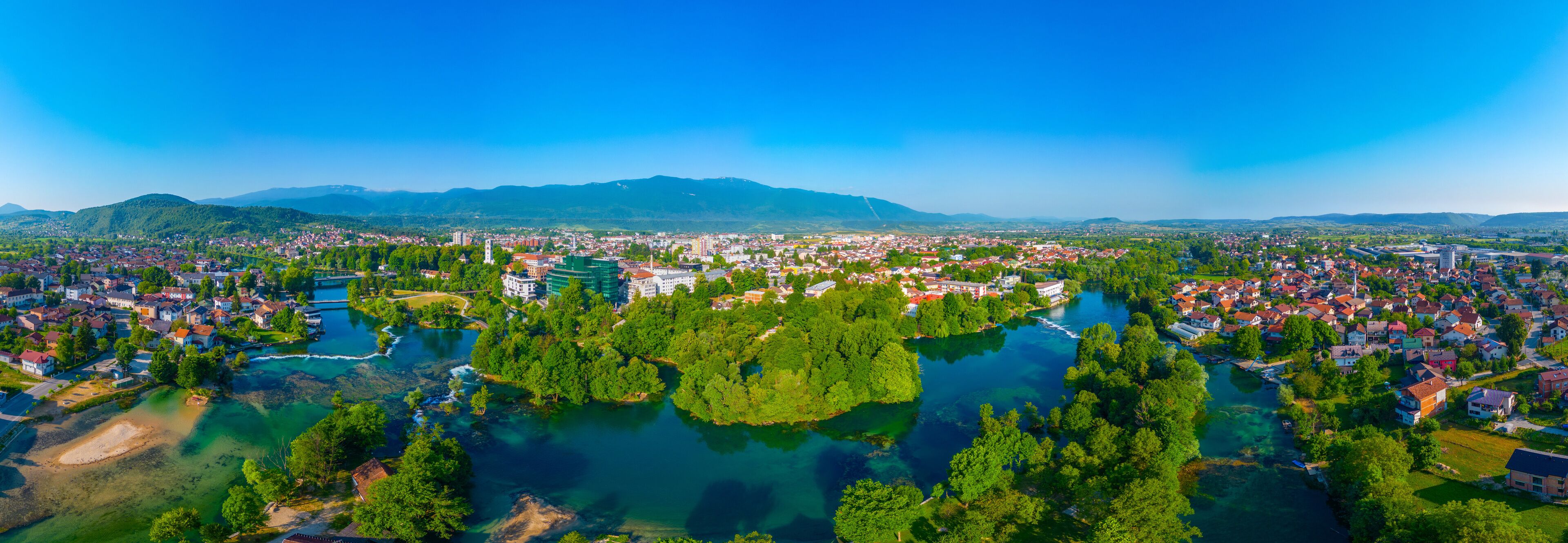 A panoramic view of a town with a river, surrounded by green hills and mountains.