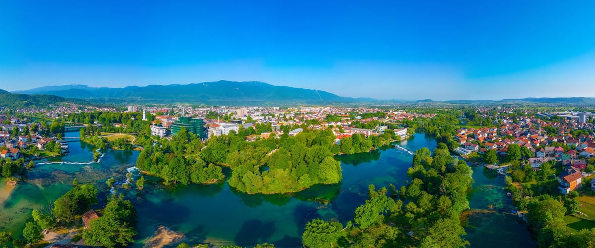 A panoramic view of a town with a river, surrounded by green hills and mountains.