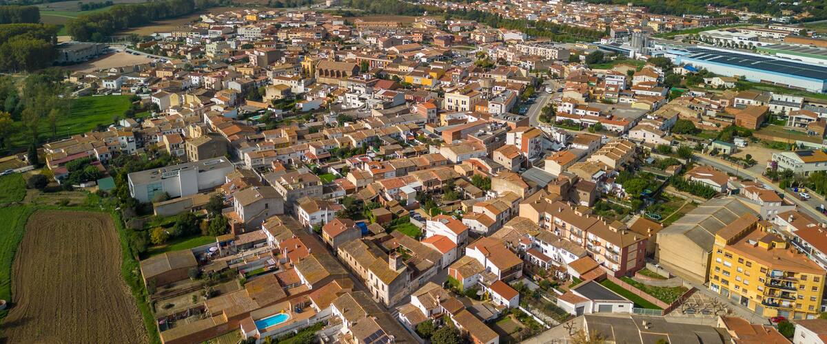 An aerial view of a densely populated town with a mix of residential and industrial buildings, surrounded by green fields and distant mountains.