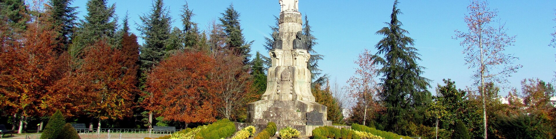Um monumento rodeado por um jardim com uma fonte, árvores com folhagem de outono e um céu azul claro.