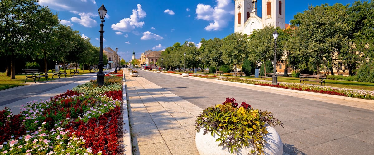 A wide boulevard with flower beds, benches, and a prominent church with twin towers.