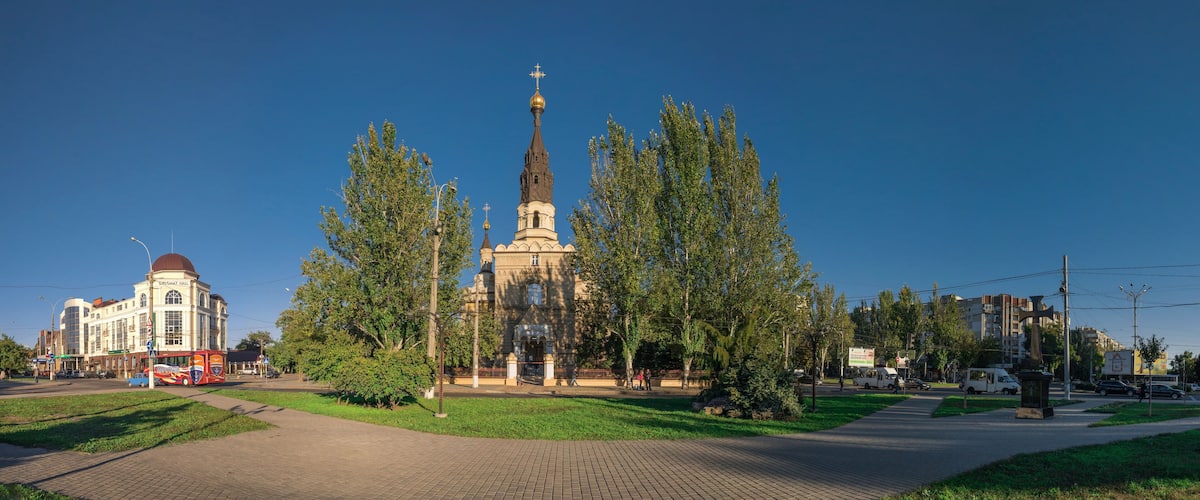 A church with a tall spire and a dome, surrounded by trees and a paved area with a bus and buildings in the background.