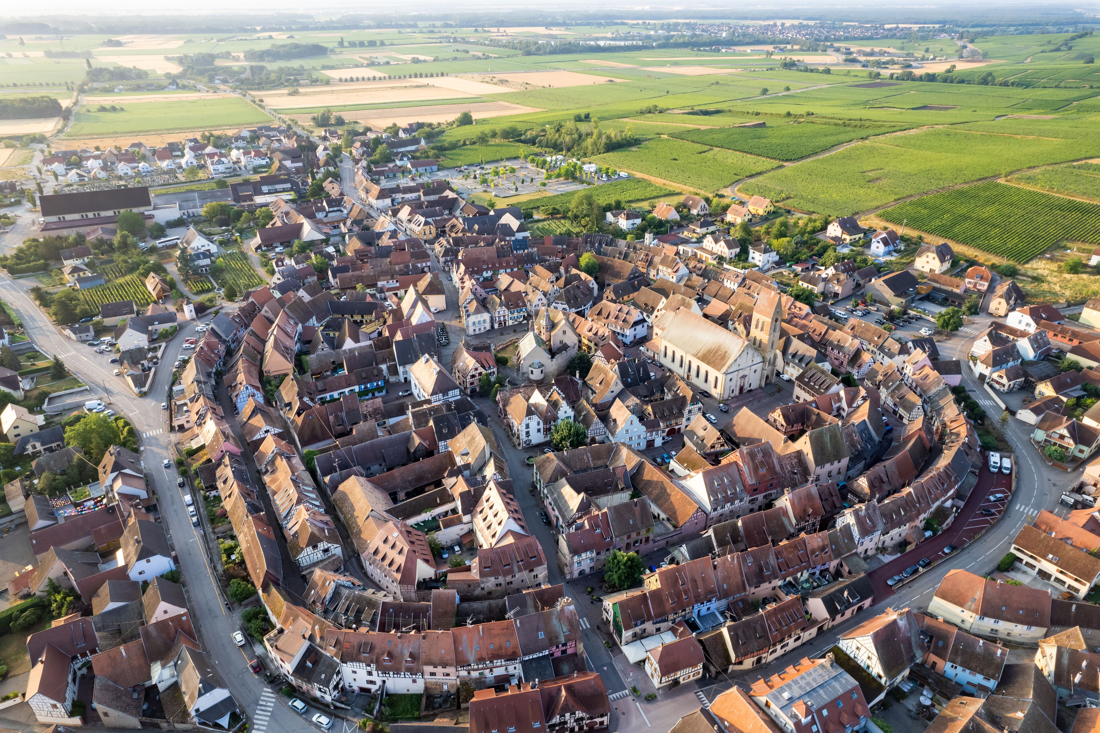 An aerial view of a historic town with a prominent church, narrow winding streets, and surrounding agricultural fields.
