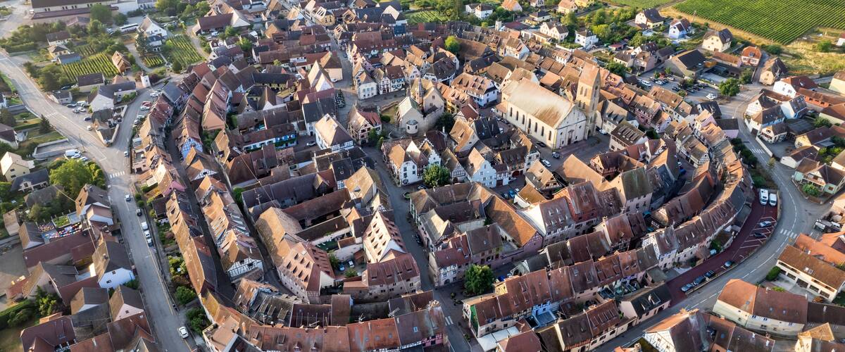 An aerial view of a historic town with a prominent church, narrow winding streets, and surrounding agricultural fields.