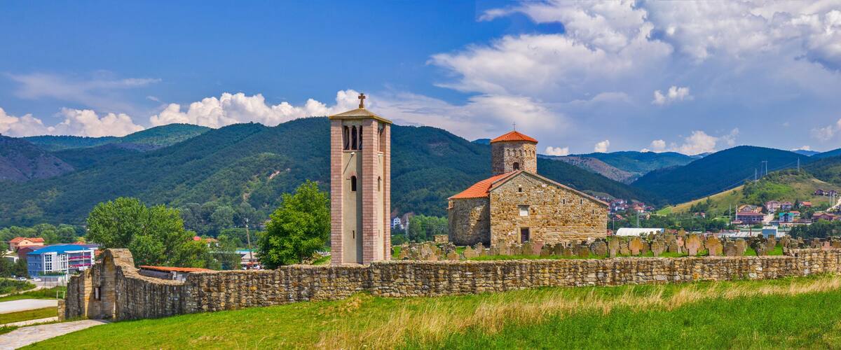 A church with a bell tower and a stone building, surrounded by mountains and a town in the background.
