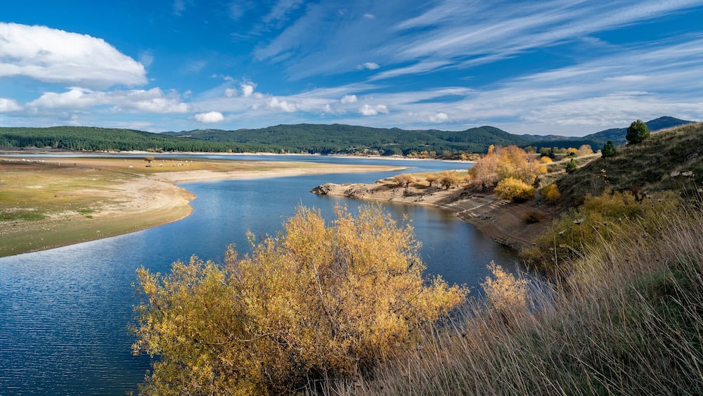 Ein Fluss schlängelt sich durch eine hügelige Landschaft mit Herbstlaub und fernen Bergen.