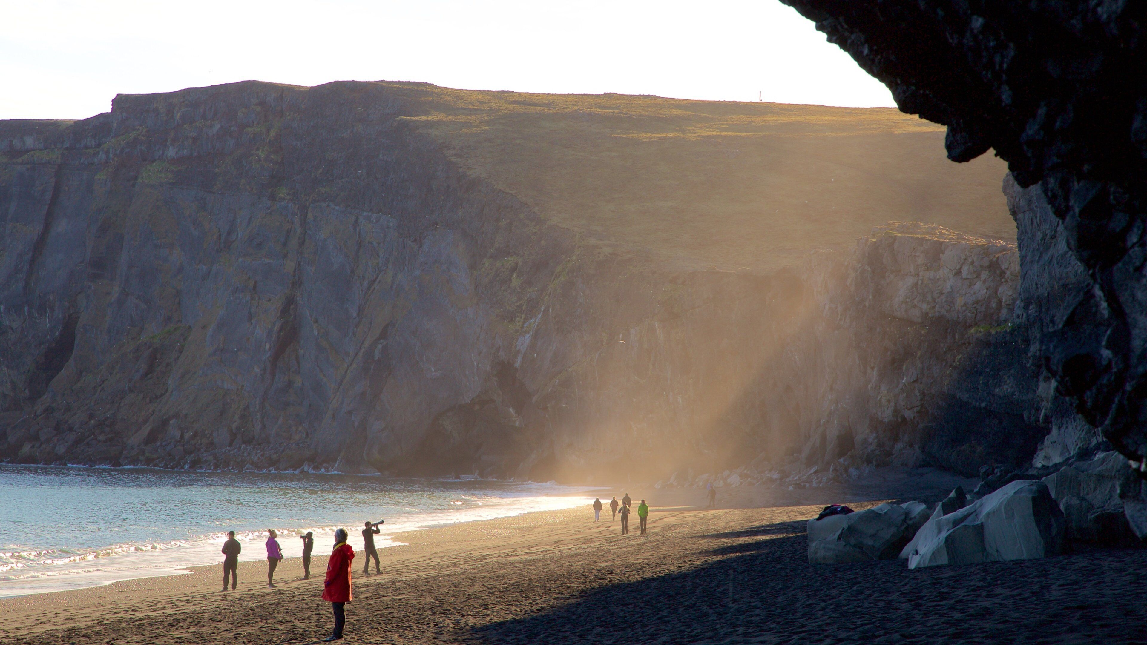 A coastal scene with a rocky cliff, a beach, and a lighthouse in the distance.
