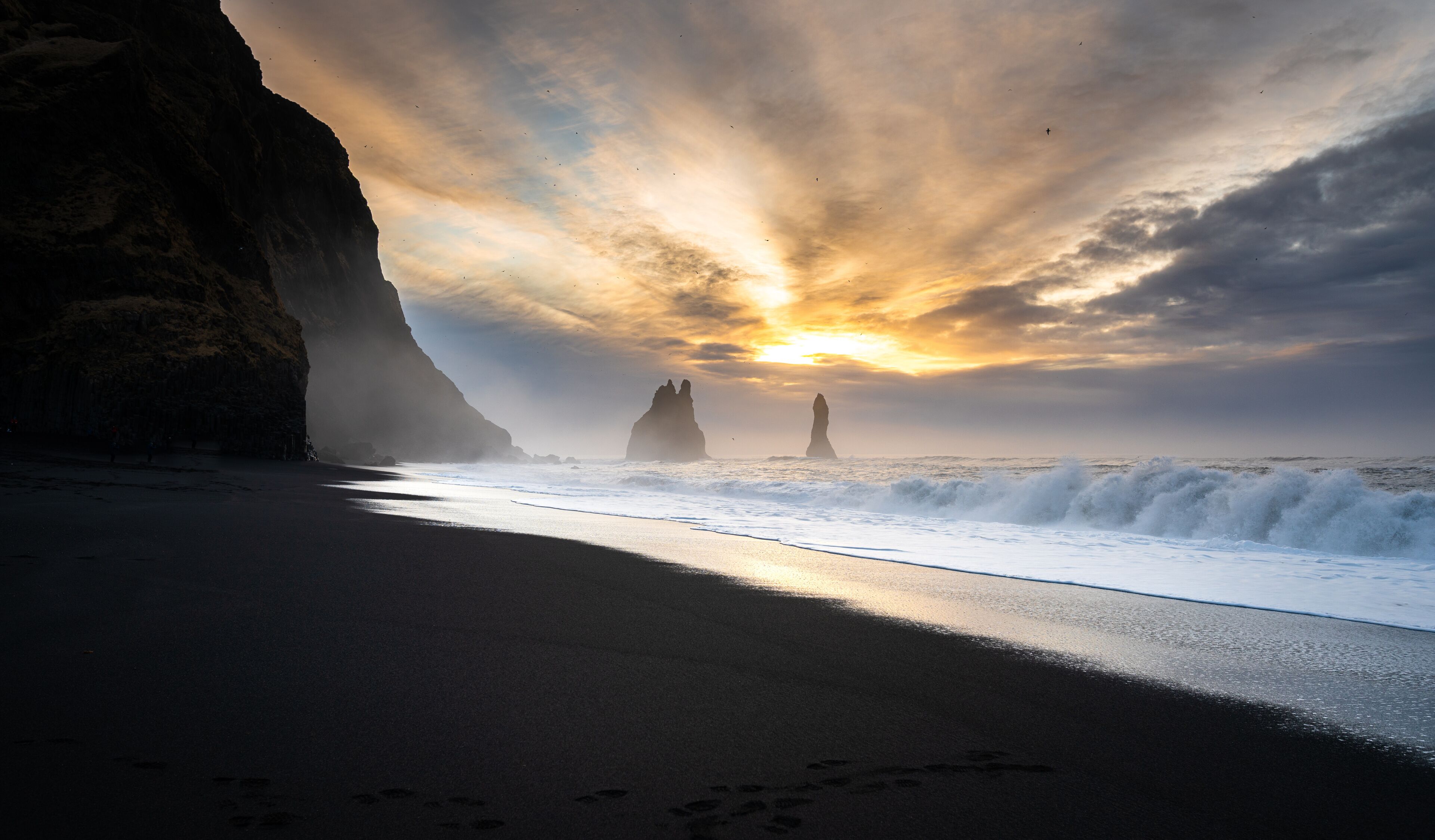A beach with waves, a cliff, and rock formations under a sunset sky.