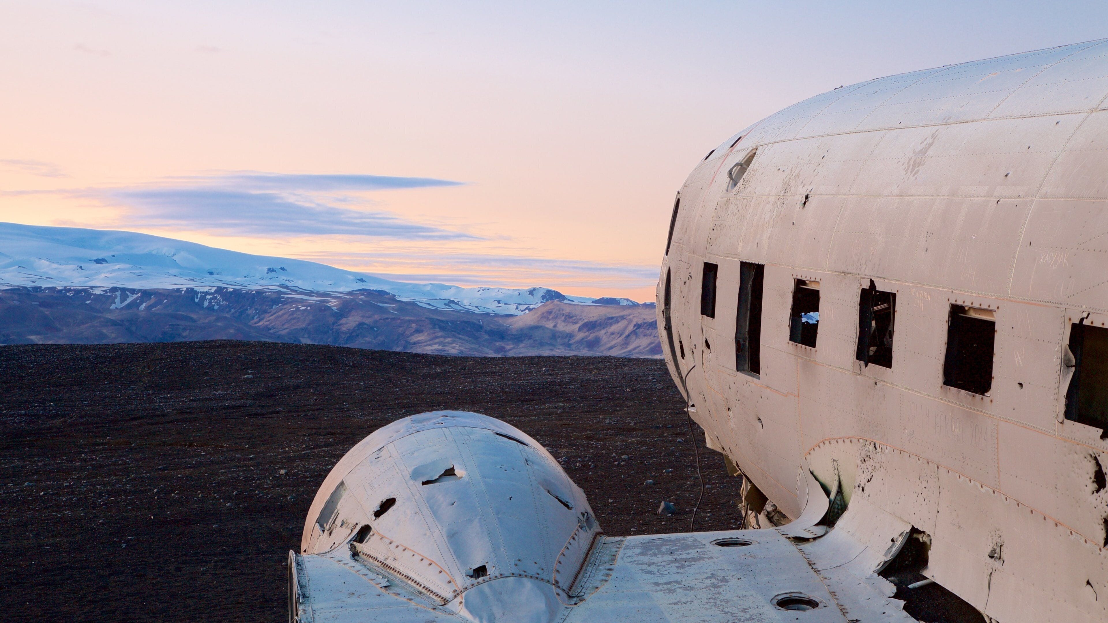 A damaged airplane in a barren landscape with snow-capped mountains in the background.