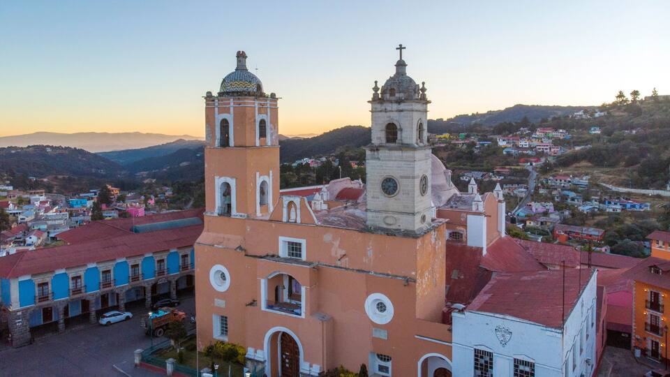 A church with two towers and a dome, surrounded by colorful buildings and a hilly landscape.