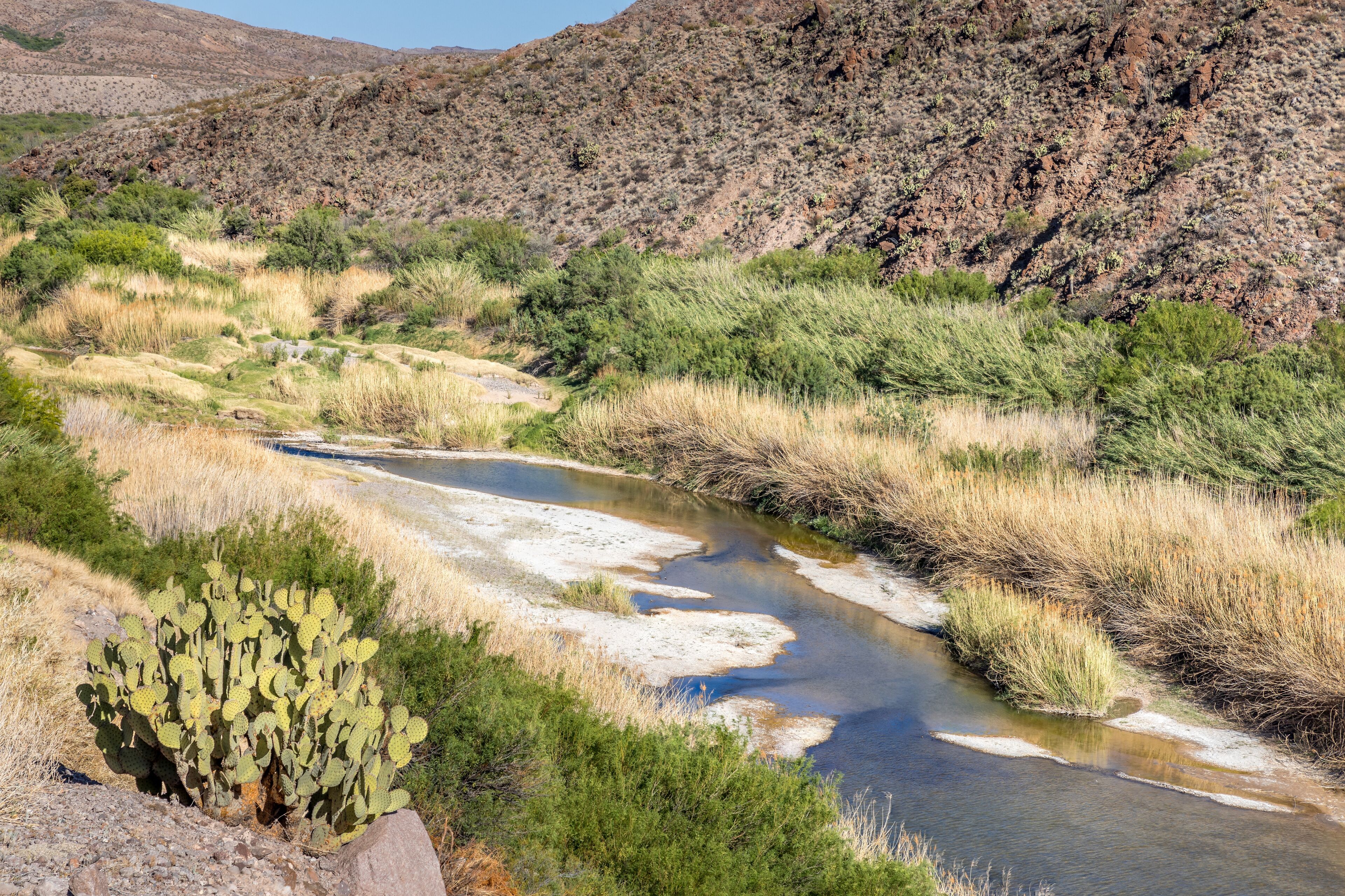 A river winding through a desert landscape with sparse vegetation and rocky hills in the background.