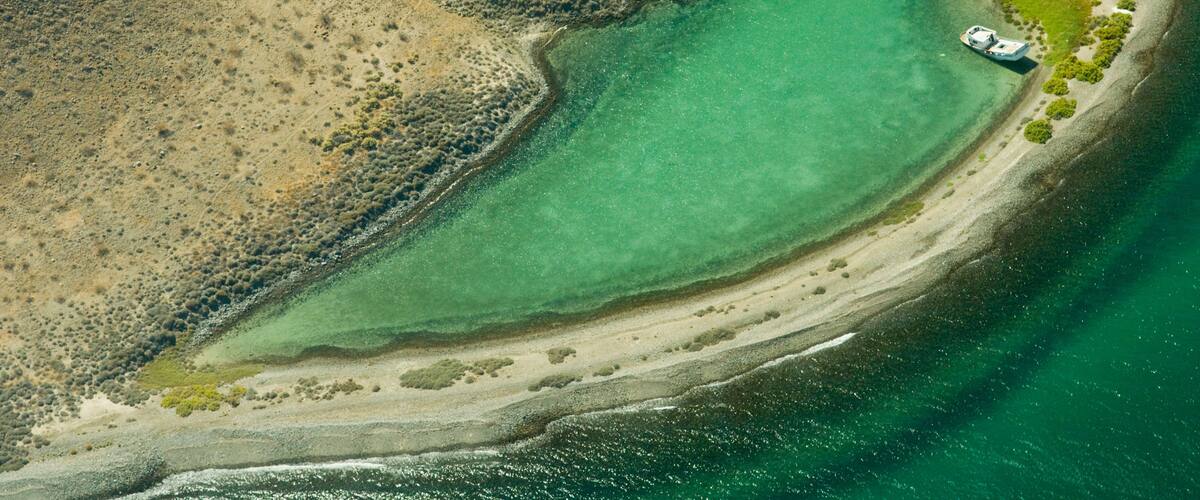 Una vista aerea di un paesaggio costiero con una piccola isola che presenta una barca, immersa in acque cristalline turchesi e spiagge sabbiose.