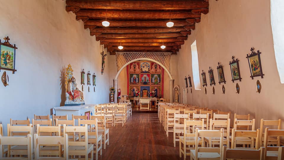 A church interior with wooden pews, a wooden ceiling, and religious artwork on the walls.