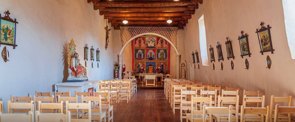 A church interior with wooden pews, a wooden ceiling, and religious artwork on the walls.