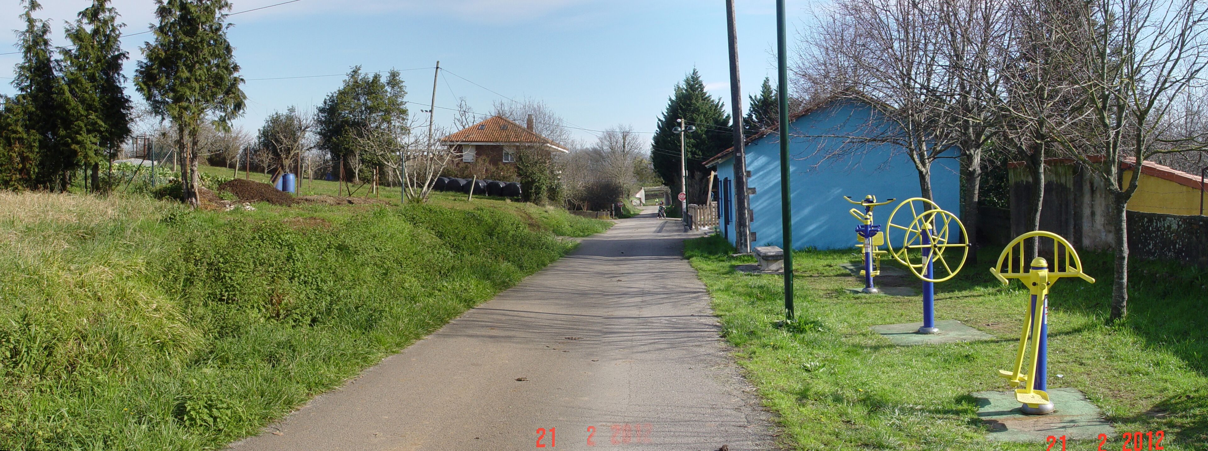 A rural pathway with a blue building, trees, and outdoor exercise equipment.